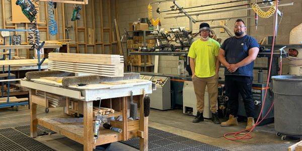 Two workers standing in a woodworking shop with equipment and wooden planks.