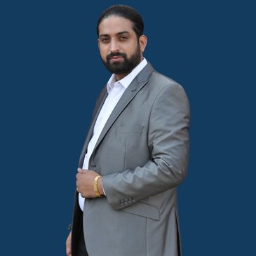 Man in a gray suit with a white shirt and gold bracelet against a blue background.