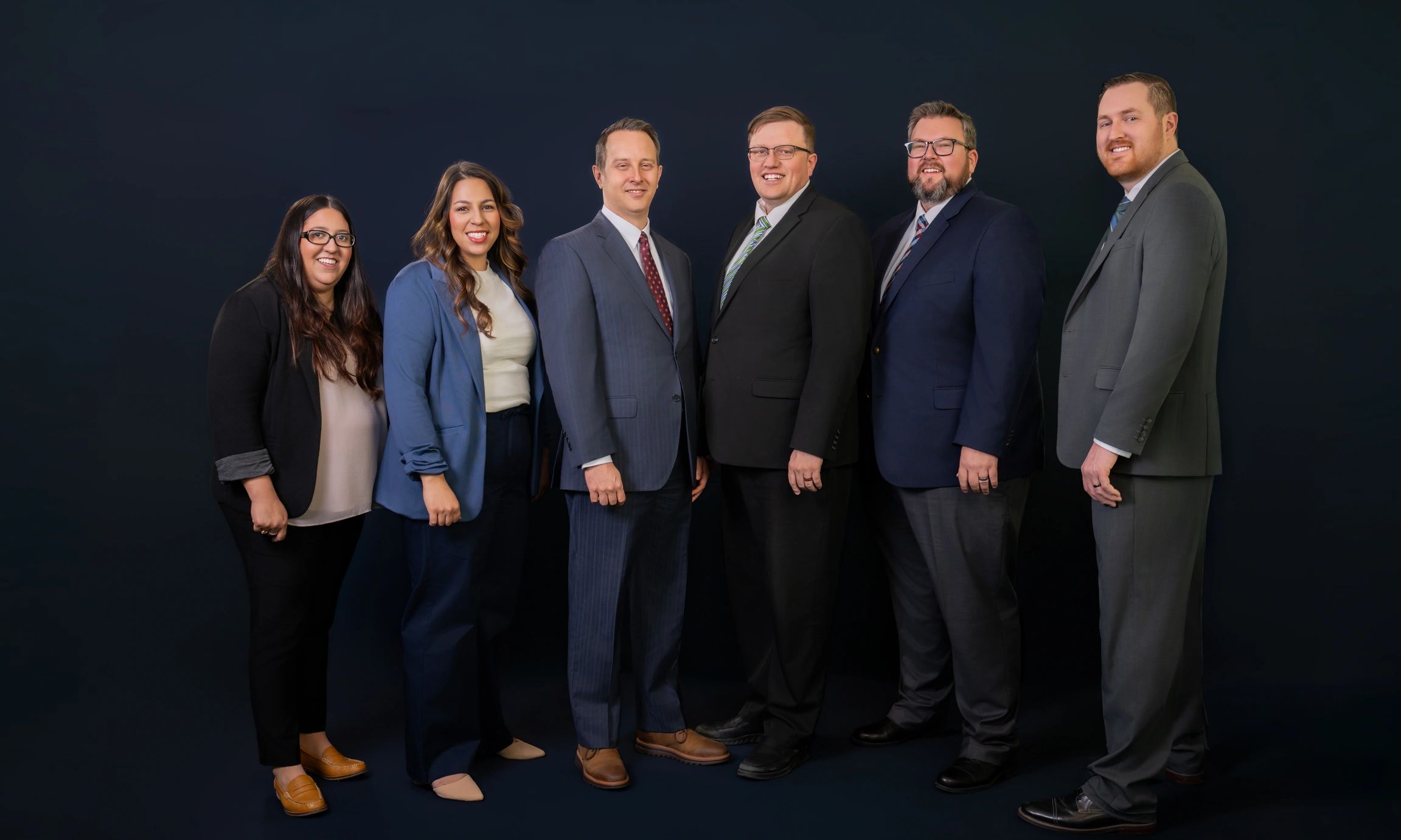 Six professionals in business attire standing against a dark background, smiling.