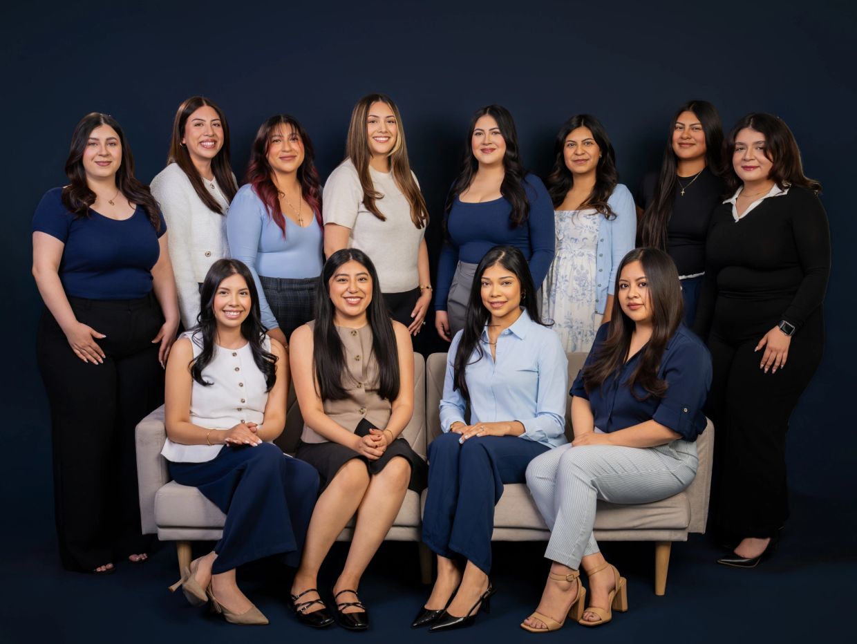 A group of 14 women posing for a formal portrait against a dark background.
