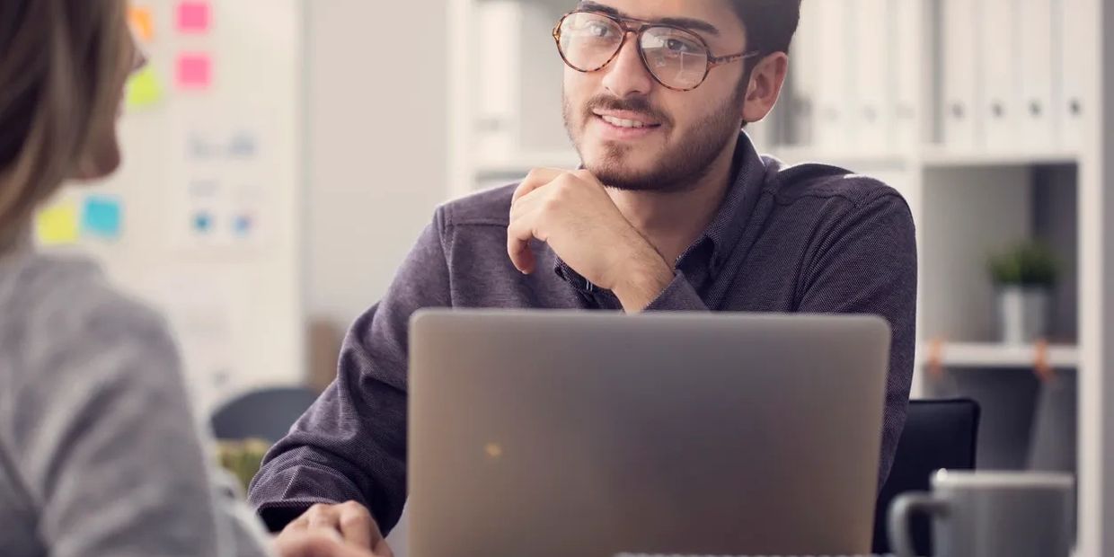 Young man with glasses working on a laptop, engaging in conversation.