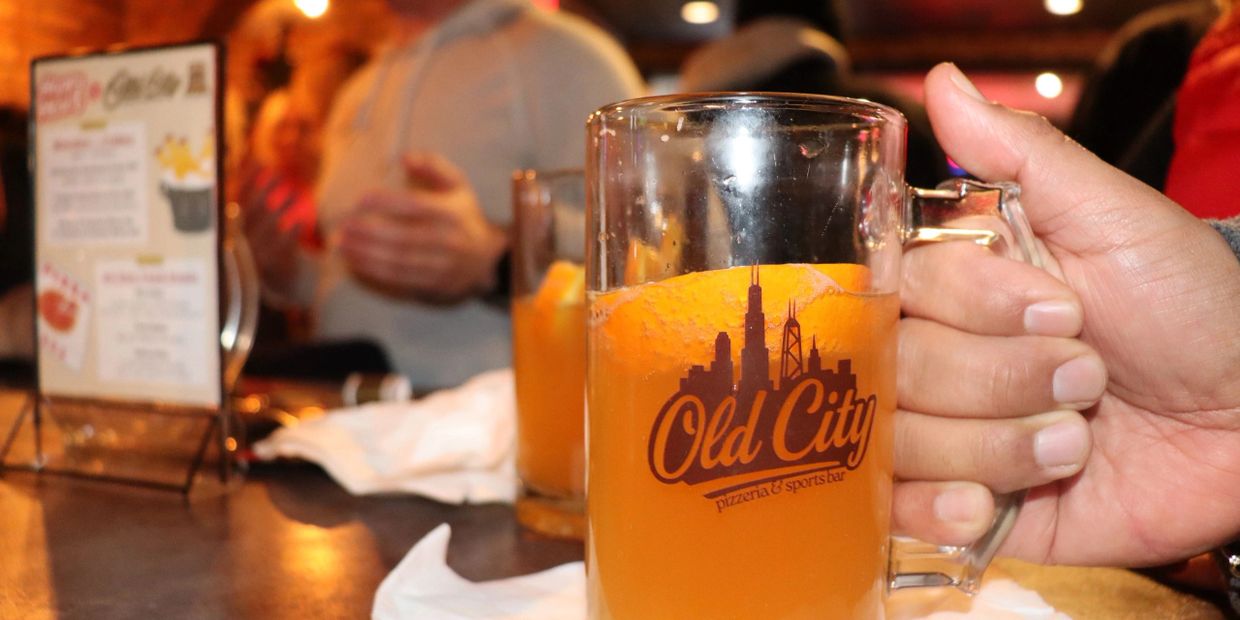 Hand holding a beer mug with 'Old City' logo in a lively bar setting.