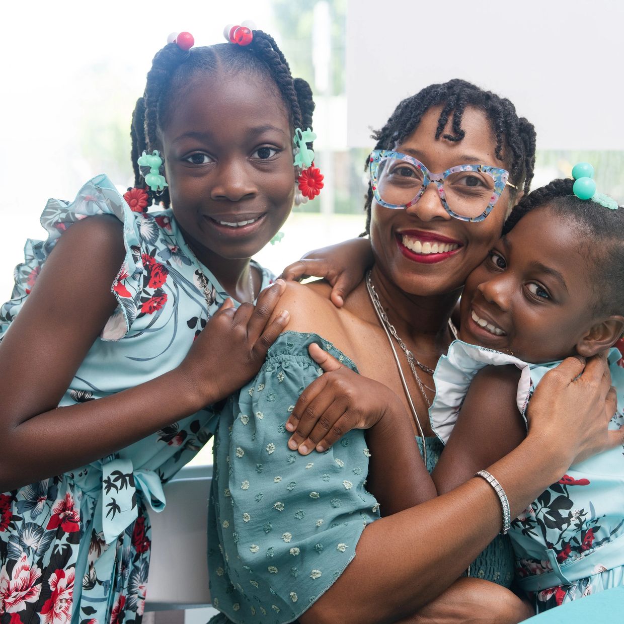 A joyful woman embraces two smiling girls in floral dresses.