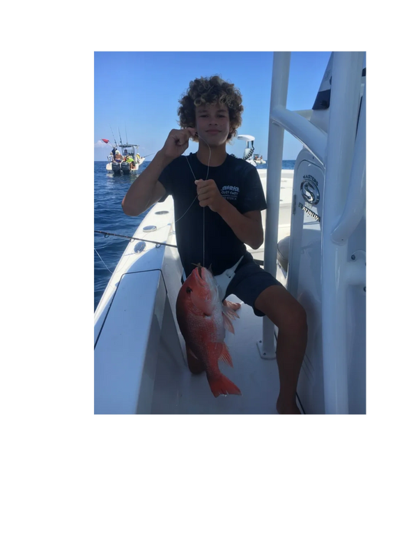 A young boy proudly holds a large red fish on a boat under clear blue skies.
