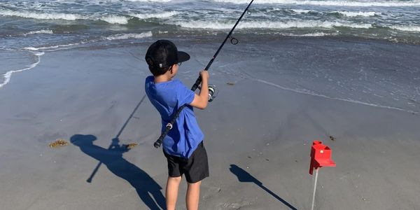 A boy fishing barefoot on a sandy beach with waves in the background.