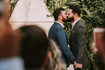 Two men pictured kissing during wedding ceremony with green foliage backdrop 