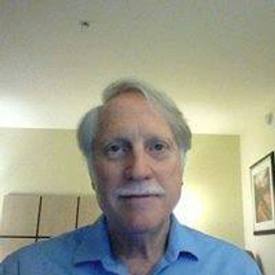 Elderly man with white hair and mustache wearing a blue shirt indoors.