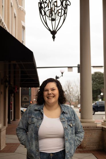 A smiling woman in a denim jacket stands on a city sidewalk under a decorative light fixture.