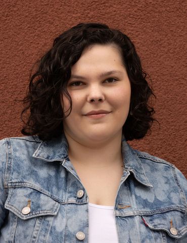 Young woman with curly dark hair wearing a denim jacket against a brick wall.