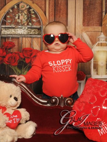 Baby in heart-shaped sunglasses and red outfit with 'Sloppy Kisser' text, surrounded by Valentine's decor.