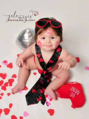 Adorable baby with heart-themed tie and sunglasses, surrounded by Valentine decorations.