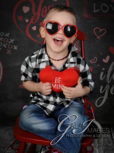 Smiling boy with heart-shaped sunglasses holding a red plush heart.