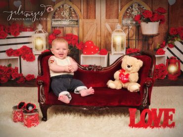 Smiling baby sitting on red velvet couch with teddy bear and love decorations.
