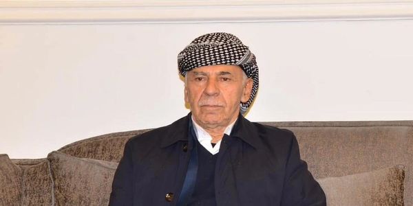 Elderly man wearing a patterned headscarf sitting on a couch.