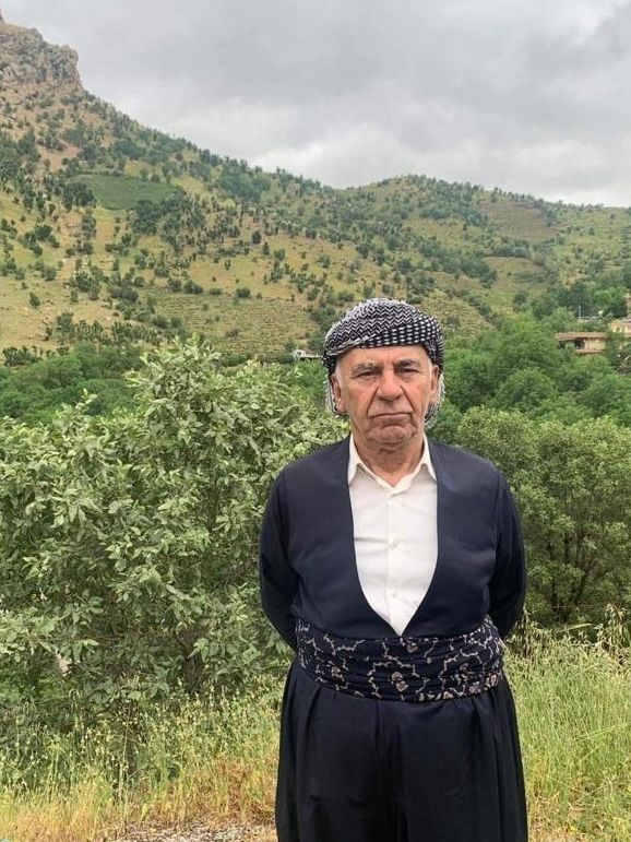 An elderly man in traditional Kurdish attire stands in a lush green mountainous area.
