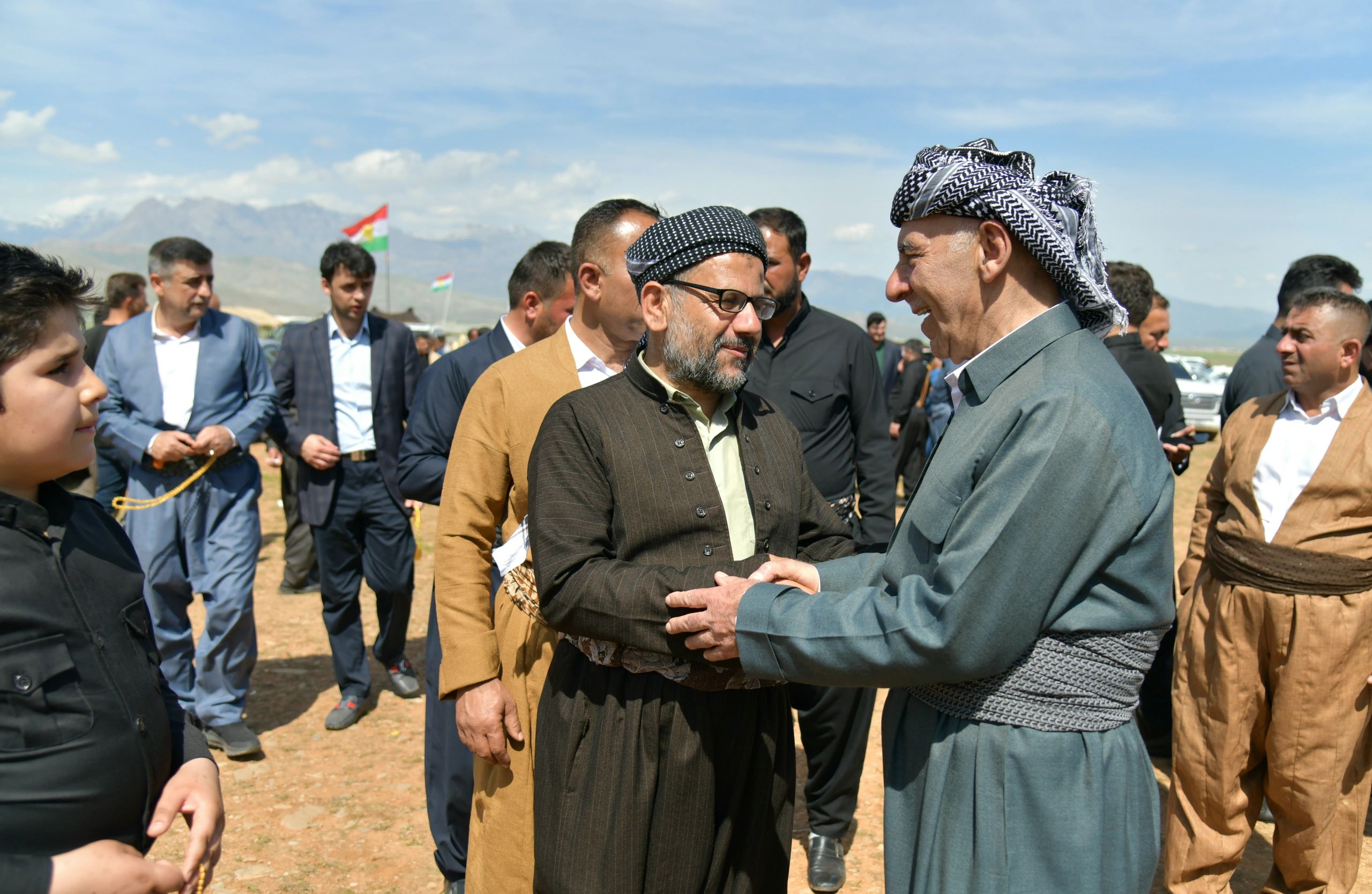 Two men wearing traditional Kurdish attire warmly greet each other outdoors.