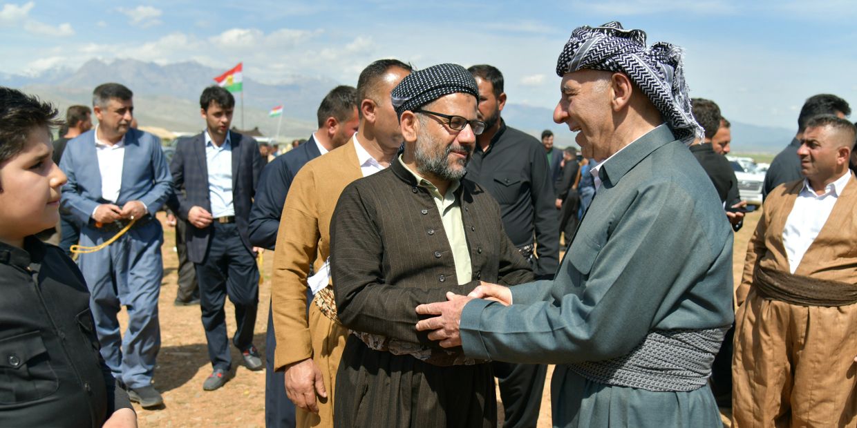 Two men in traditional Kurdish attire warmly greet each other outdoors with others around them.