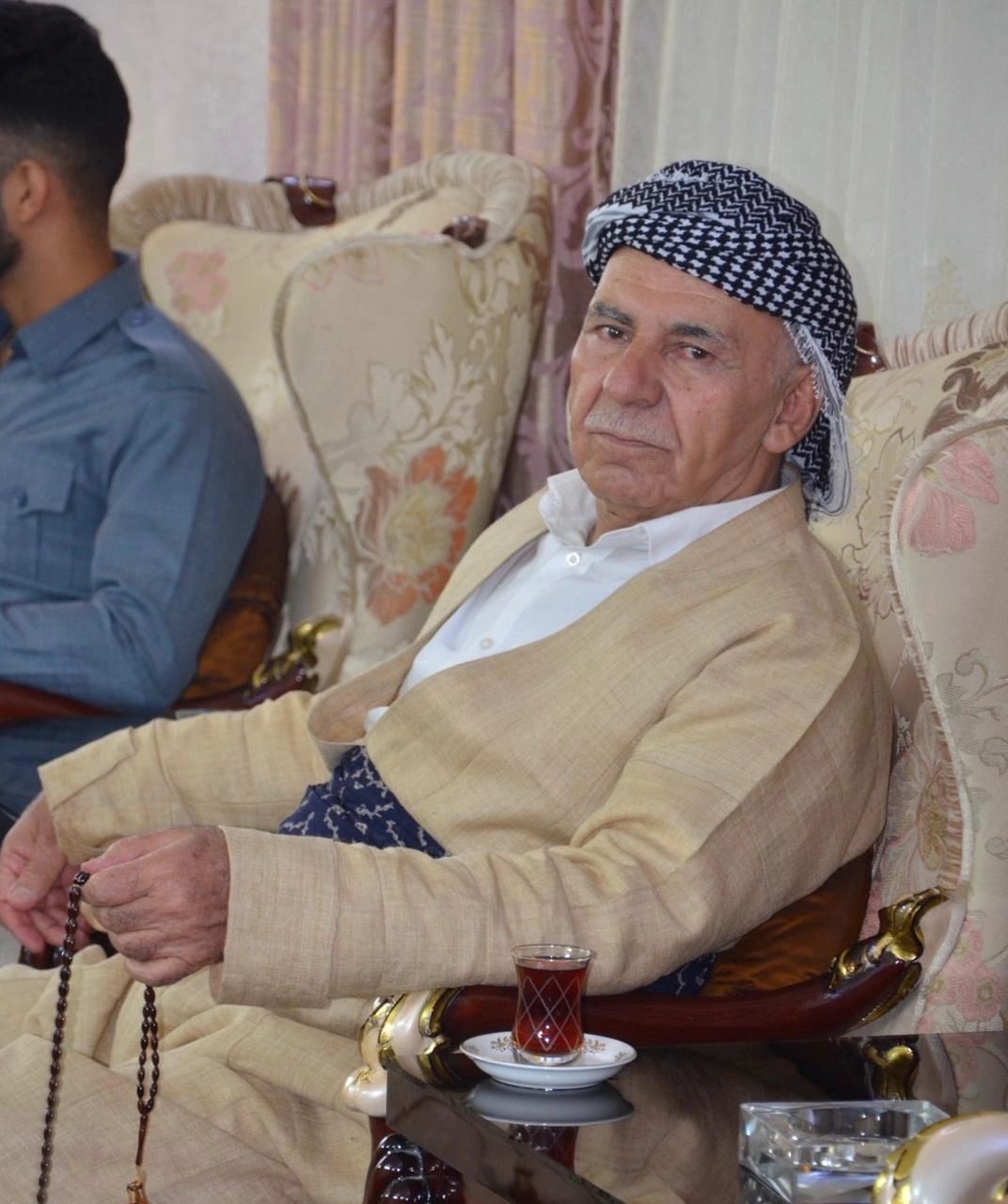 Elderly man in traditional attire holding prayer beads, sitting on an ornate chair with tea.