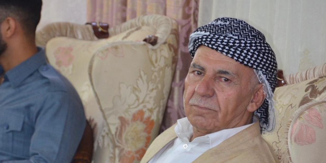 Elderly man wearing traditional headscarf, sitting on a floral-patterned sofa.