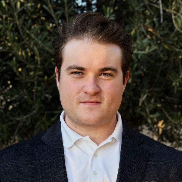 A young man in a white shirt and dark blazer stands outdoors with greenery behind him.
