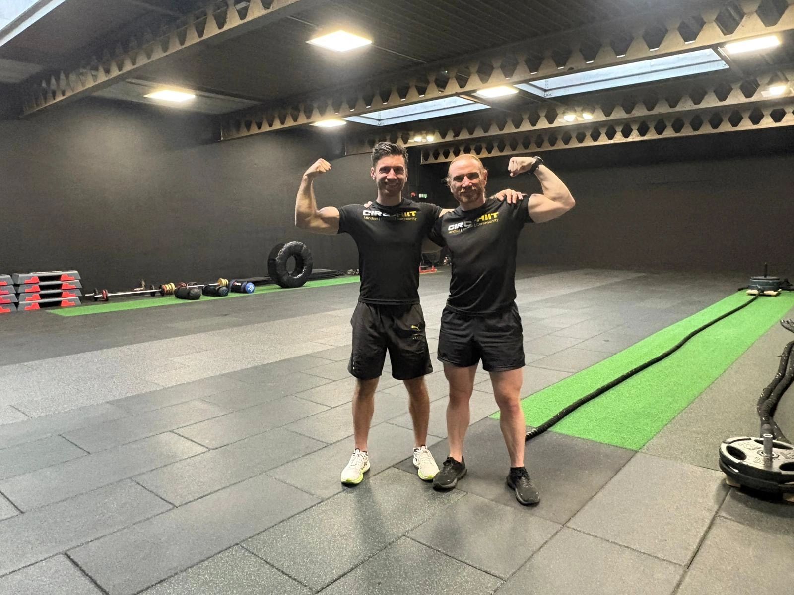 Two men flexing muscles in a gym with workout equipment around.