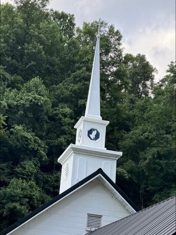 White church steeple with a cross amidst green trees on a cloudy day.