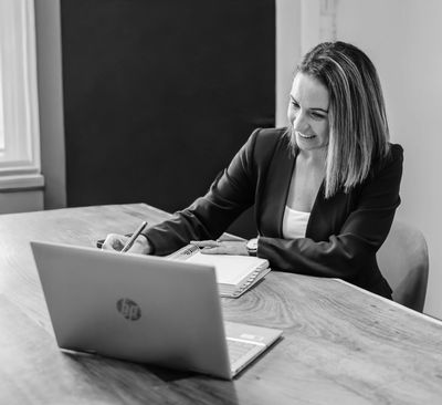 Smiling woman in business attire writing notes during a video call on a laptop.
