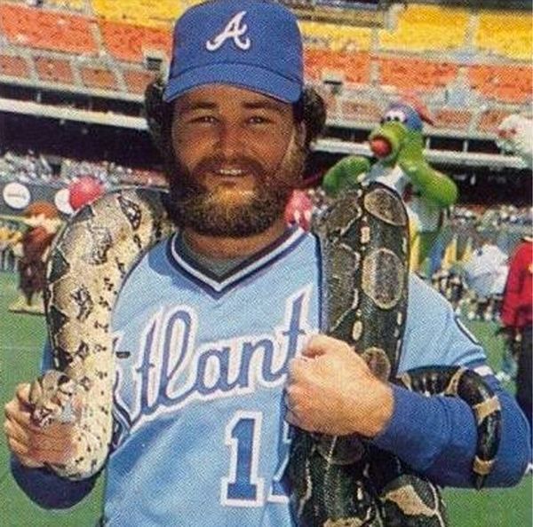 Man in Atlanta baseball uniform holding two large snakes at a stadium.