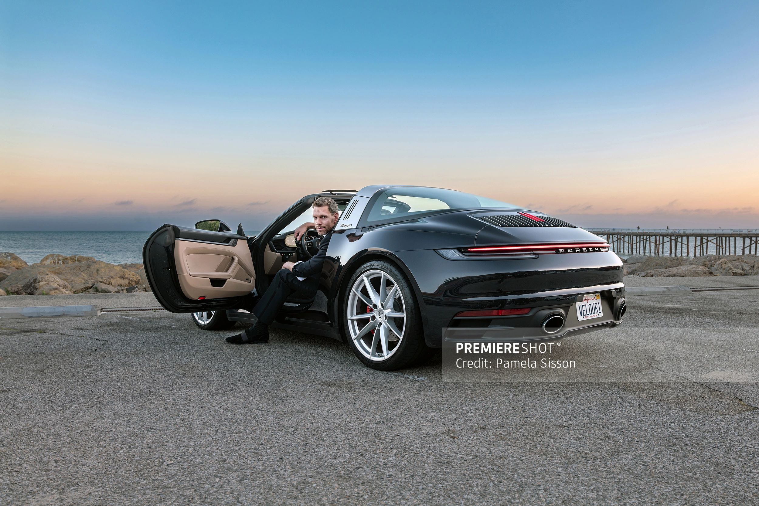 Man sitting in a black Porsche Targa with ocean and pier in the background.
