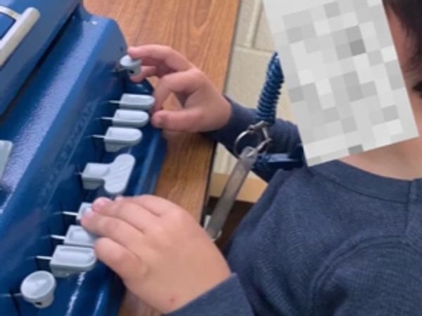 PD: A young boy is pictured writing on a blue Perkins braille writer.