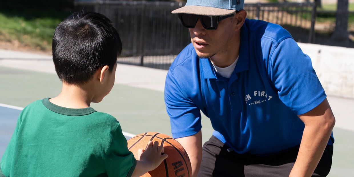 A man coaches a young boy playing basketball outdoors on a sunny day.