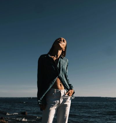 A woman enjoys the sun by the ocean, dressed in a loose shirt and white pants.