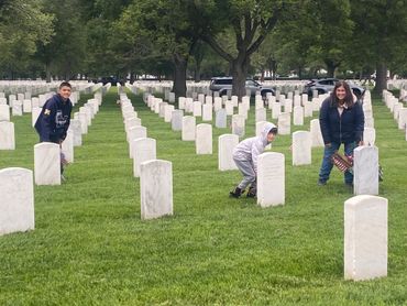 Three people visiting a cemetery with rows of white headstones on green grass.