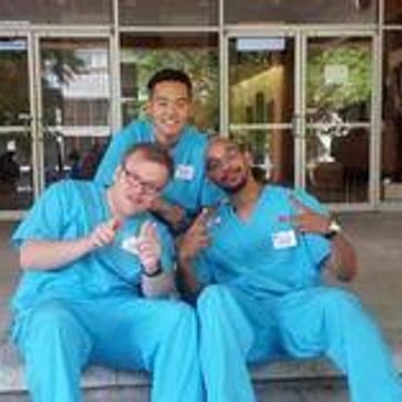 Three medical professionals in blue scrubs posing and smiling outside a building.