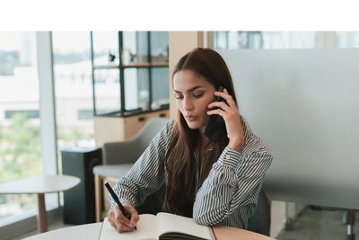 Young woman multitasking by talking on phone and writing in notebook.