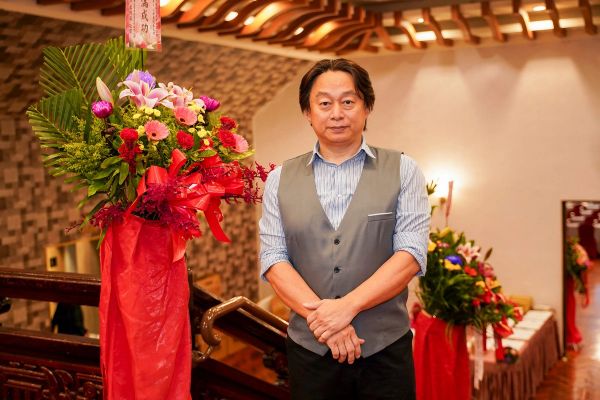 Man in a vest standing beside a large floral arrangement indoors.