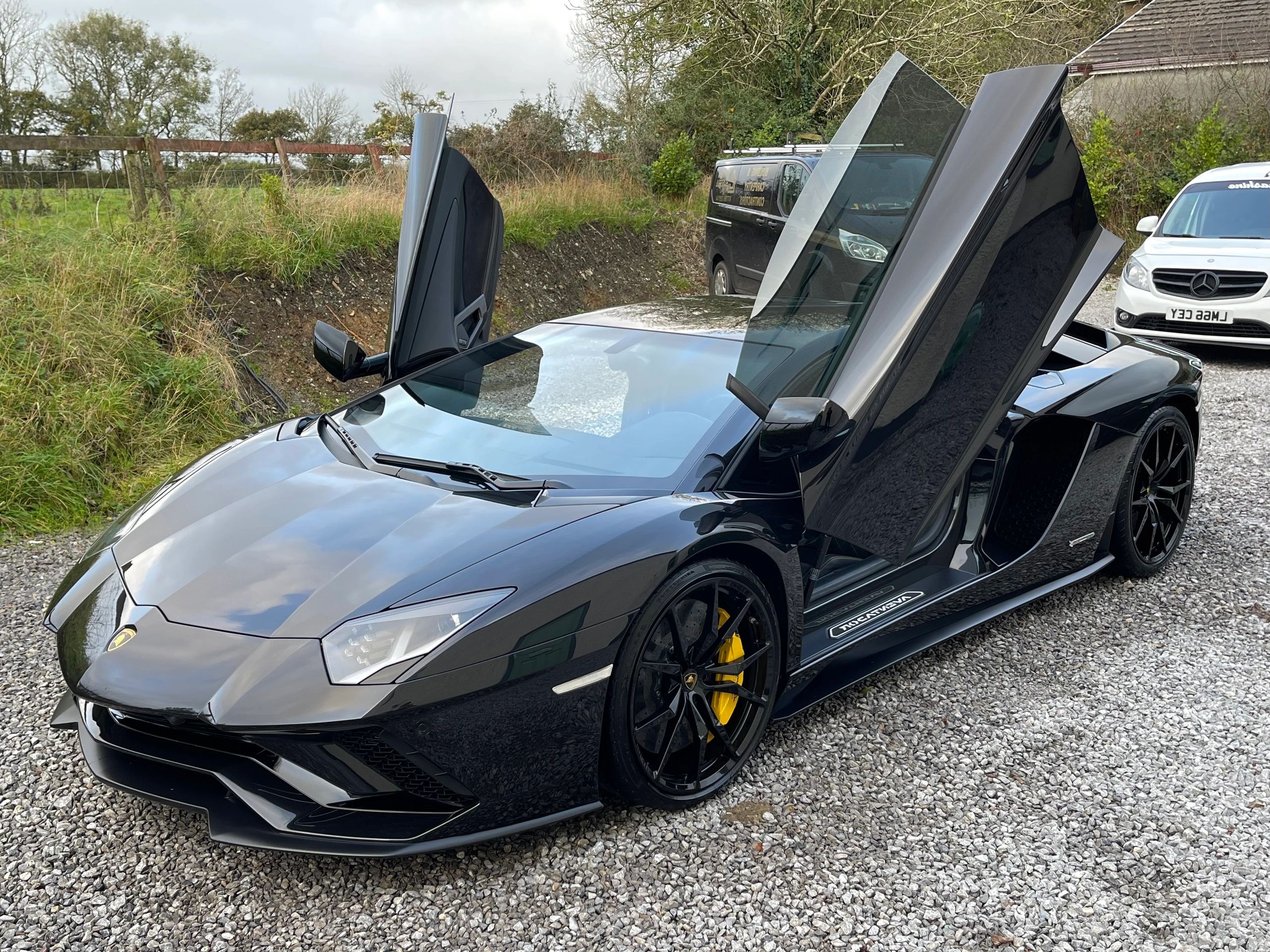 Black Lamborghini Aventador with scissor doors open, parked on gravel.