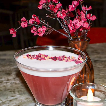 Elegant pink cocktail garnished with rose petals, with a small candle and vase of red flowers.
