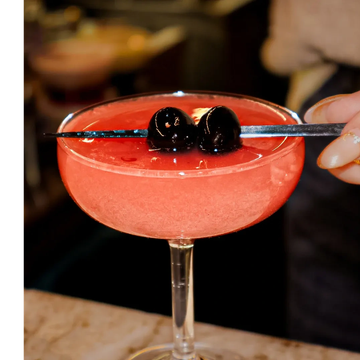 A pink cocktail garnished with two dark cherries on a skewer.