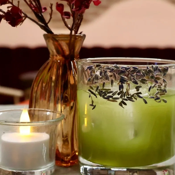 A lit candle and a green lavender-scented candle beside a brown vase with dried flowers.