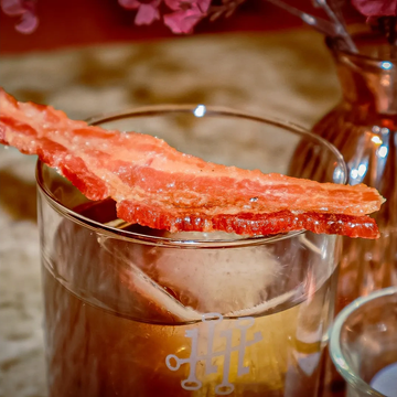 Cocktail garnished with a crispy bacon strip and ice cube.