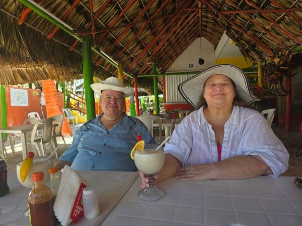 Two people wearing hats sitting at a table under a thatched roof with drinks.