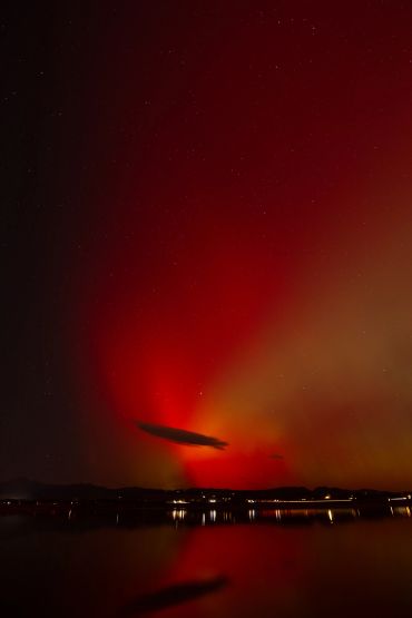 Red aurora lights up the night sky over a calm lake with silhouetted mountains.