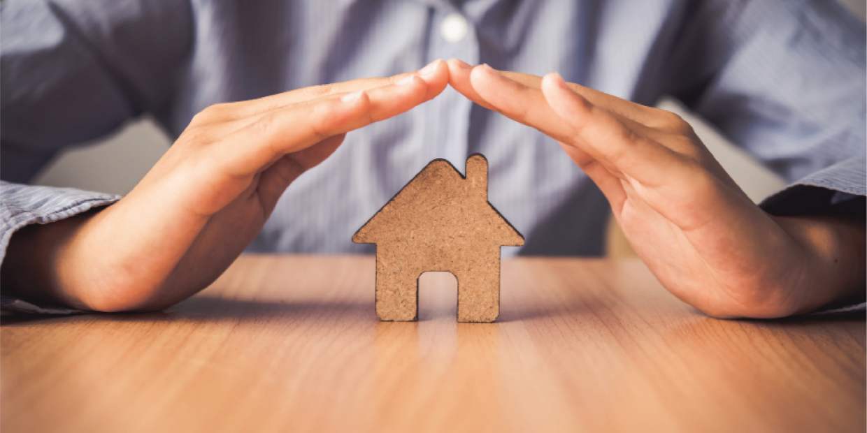 Hands protect a small wooden house model on a table.