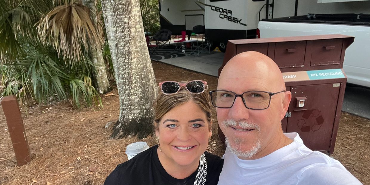 A cheerful couple takes a selfie outdoors near trees and an RV.