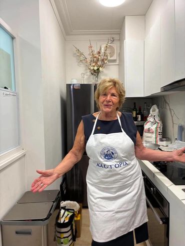 An elderly woman in a white apron stands happily in a kitchen.
