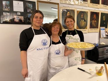 Three women in aprons posing with a tray of uncooked pasta in a kitchen.