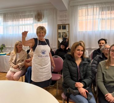 Woman in apron smiling and waving in a cozy room with seated people.