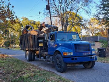 Blue truck carrying large cut logs on a suburban street in autumn.
