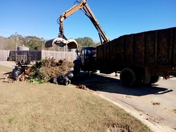 Excavator loading yard waste and black bags into a large truck under clear blue sky.